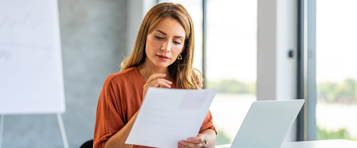 A businessperson sitting in a well-lit meeting room looks between a paper document and an open laptop, demonstrating one potential use of intelligent document processing.