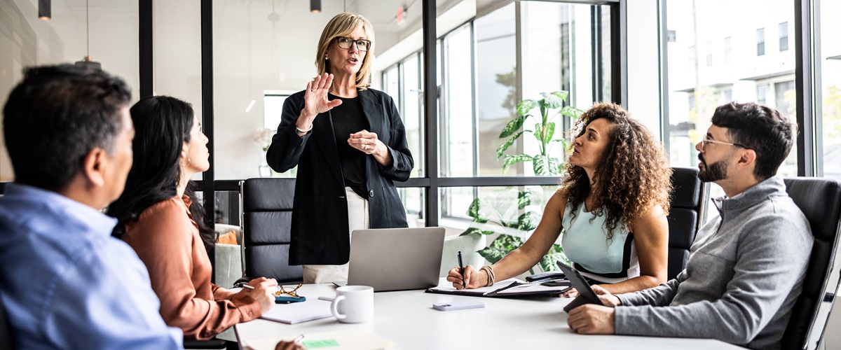 A woman leads a meeting with four coworkers in a brightly lit office.