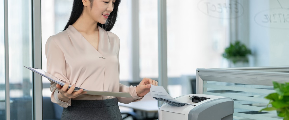 A woman lifts a sheet of paper from the top of a desktop scanner.