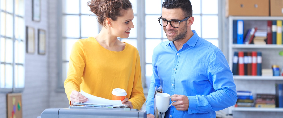 A man and a woman drink coffee and talk in front of an office printer.