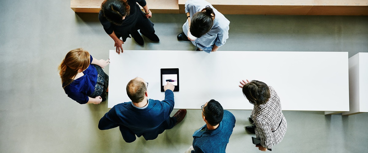 Top-down view of a group of business associates standing around a table, each one looking down toward a single document.