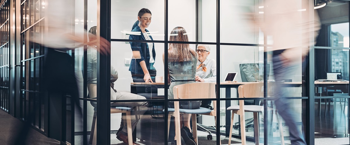 Four office workers gather inside a meeting room as other employees walk by.