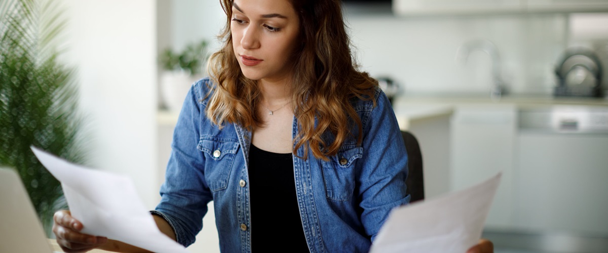 A woman analyzes a small stack of papers while sitting in front of a laptop.