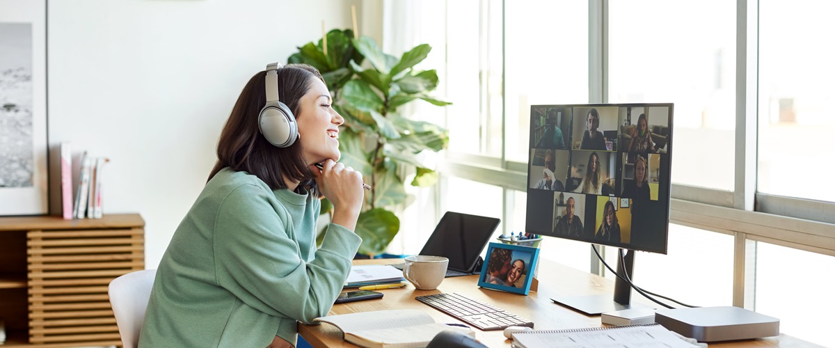 A person seated at a desk in a brightly lit work from home setup talks with colleagues through a video call.