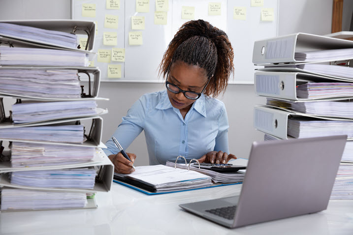 Better scanning for accounting - A woman working with a pen and a binder open at a desk surrounded by binders piled high loaded with papers.