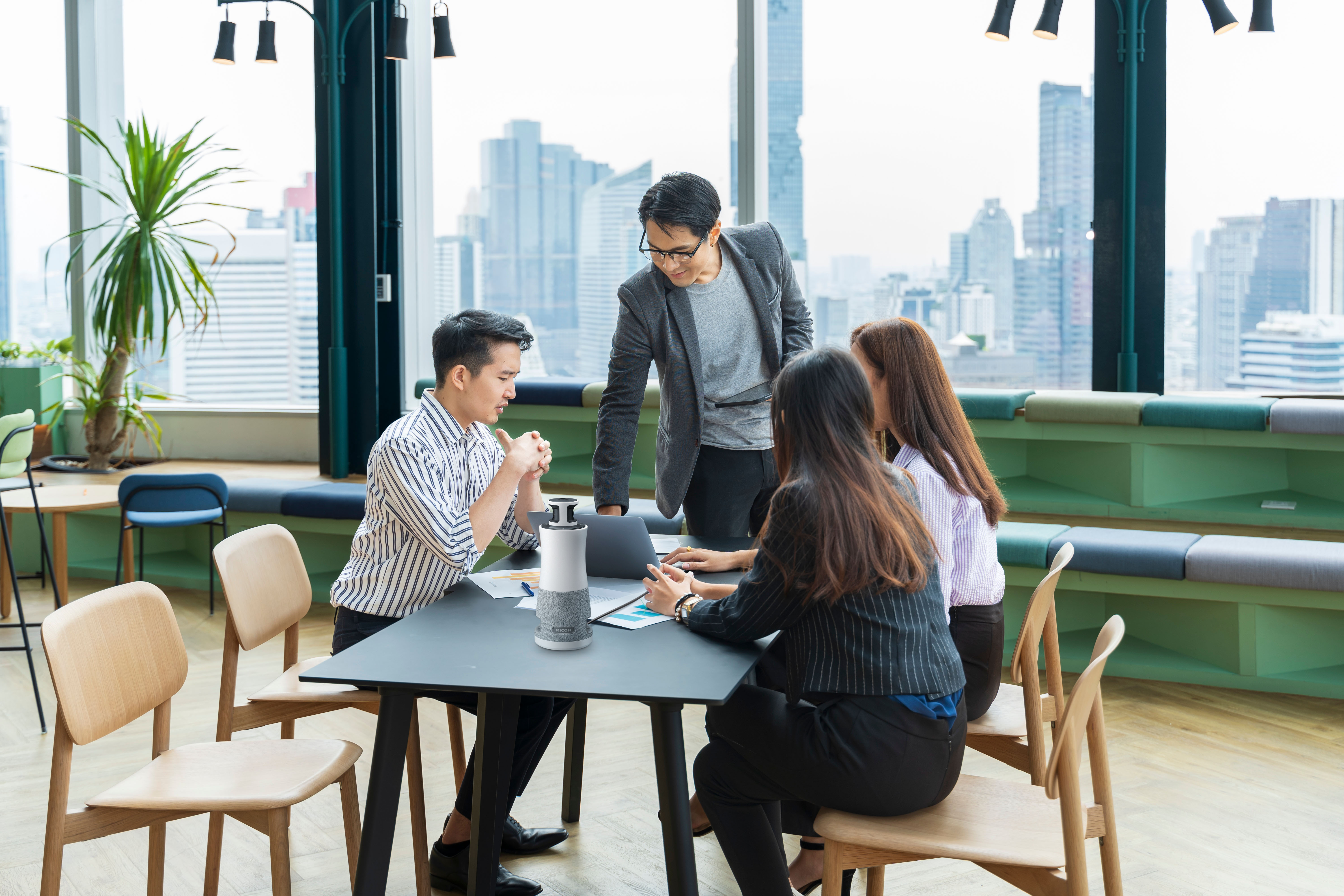 A group of four people dressed in business attire and mid discussion while seated and standing around a small rectangular table with Meeting360 in the middle of the table.